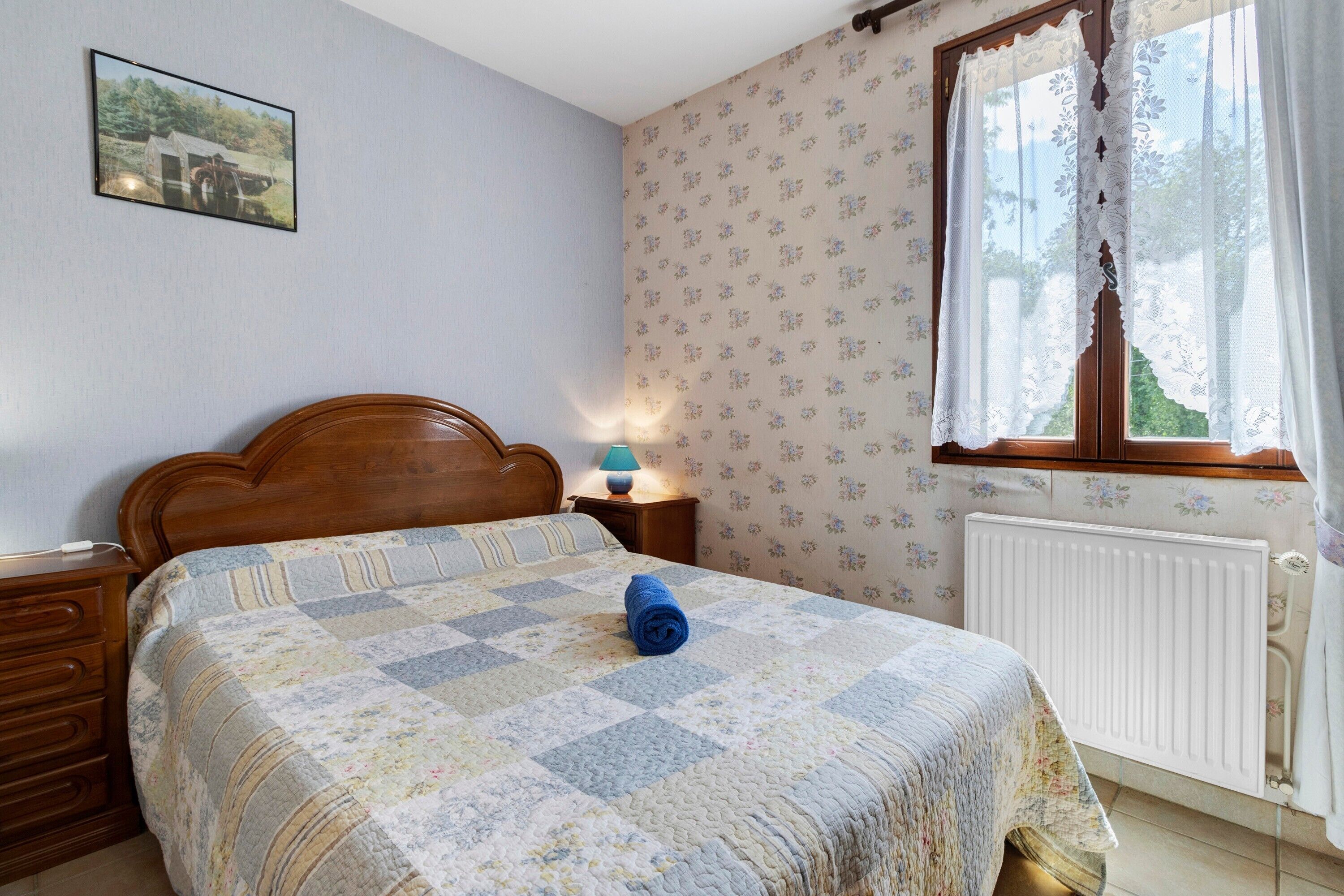 Une chambre à coucher avec un lit en bois, une courtepointe, une table de chevet avec une lampe, une fenêtre ornée de rideaux en dentelle et un radiateur.