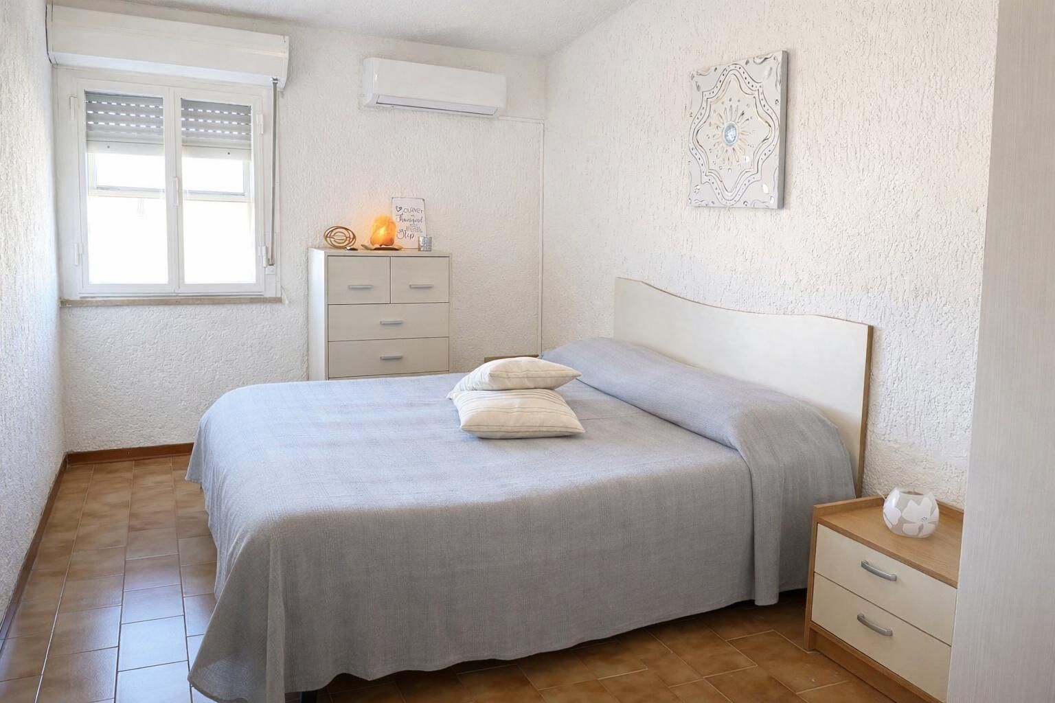 A bedroom with a bed, a nightstand, a dresser, a window, and an air conditioner.