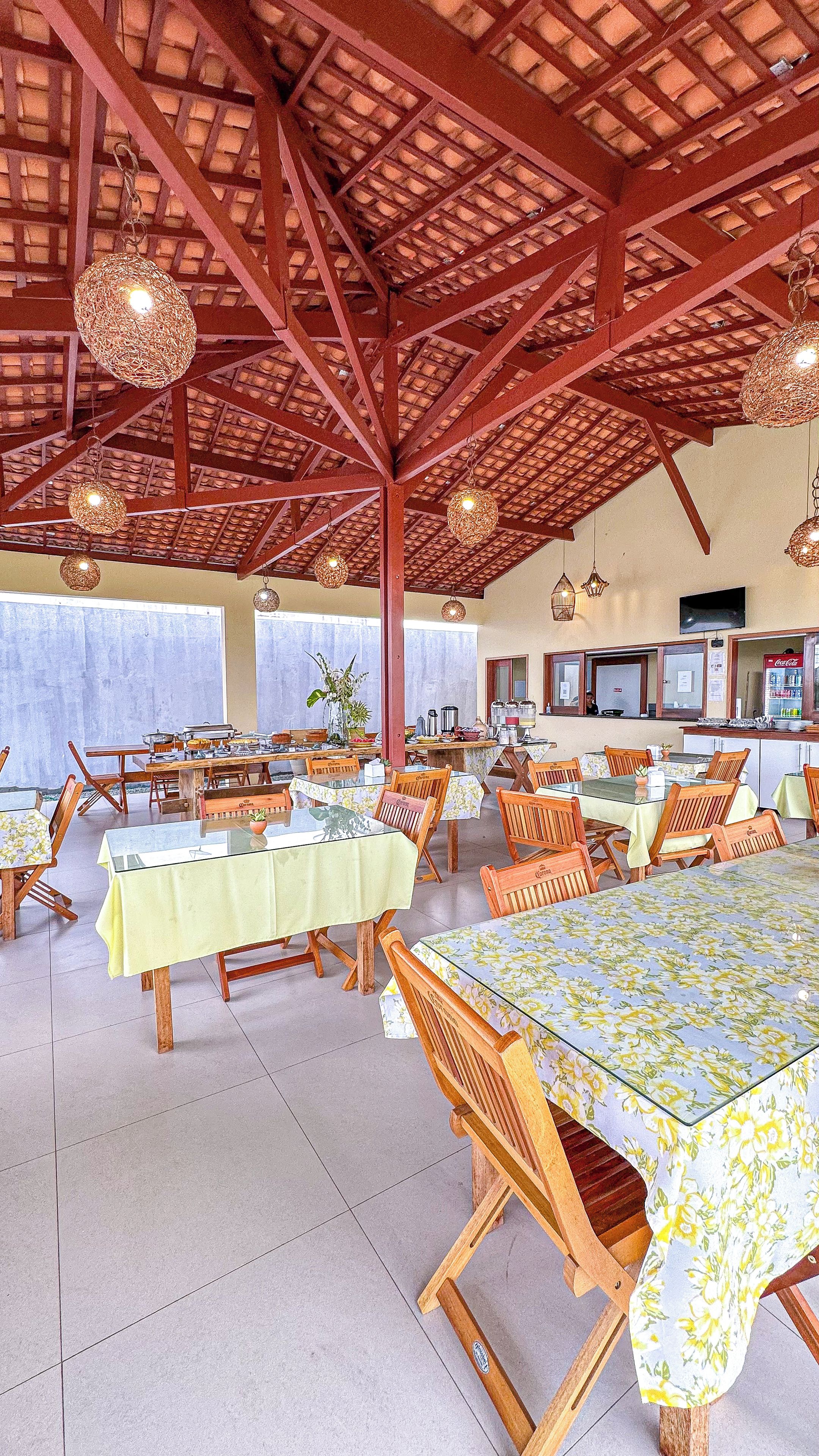 Un comedor interior con mesas y sillas, suelo de baldosas y un techo alto con vigas de madera.
