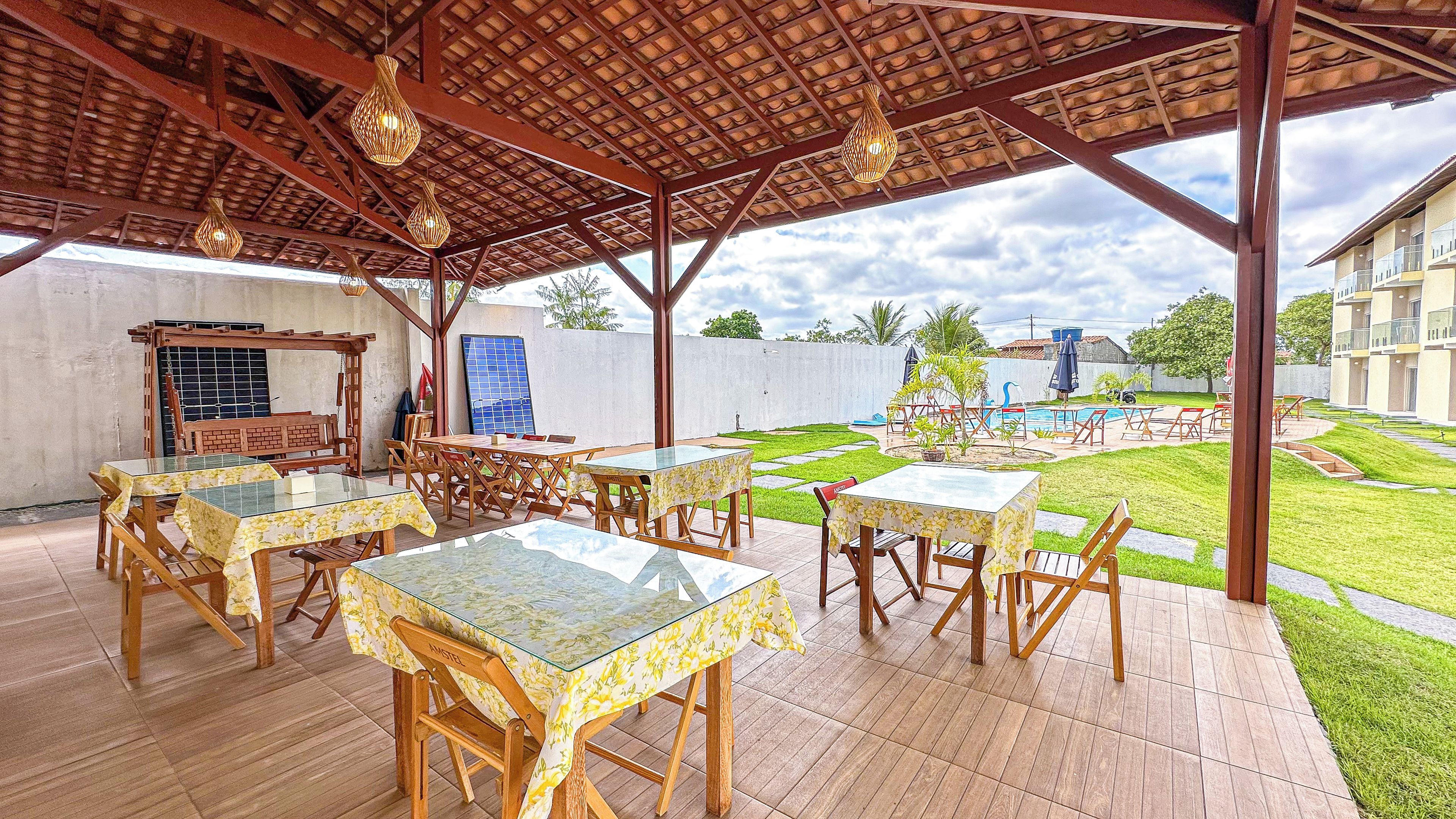Une terrasse extérieure avec des tables et des chaises en bois, une pergola décorée de guirlandes lumineuses, et une vue sur une piscine et des bâtiments en arrière-plan.