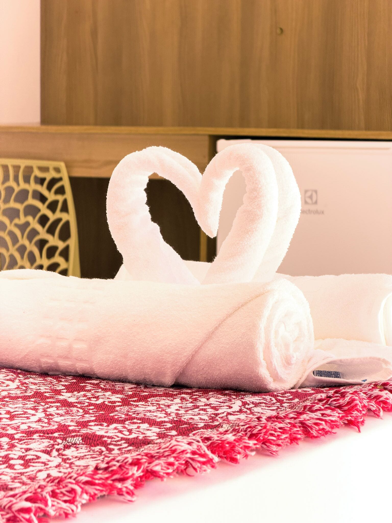 A rolled white towel with a heart shape on a bed with a red patterned bedspread.