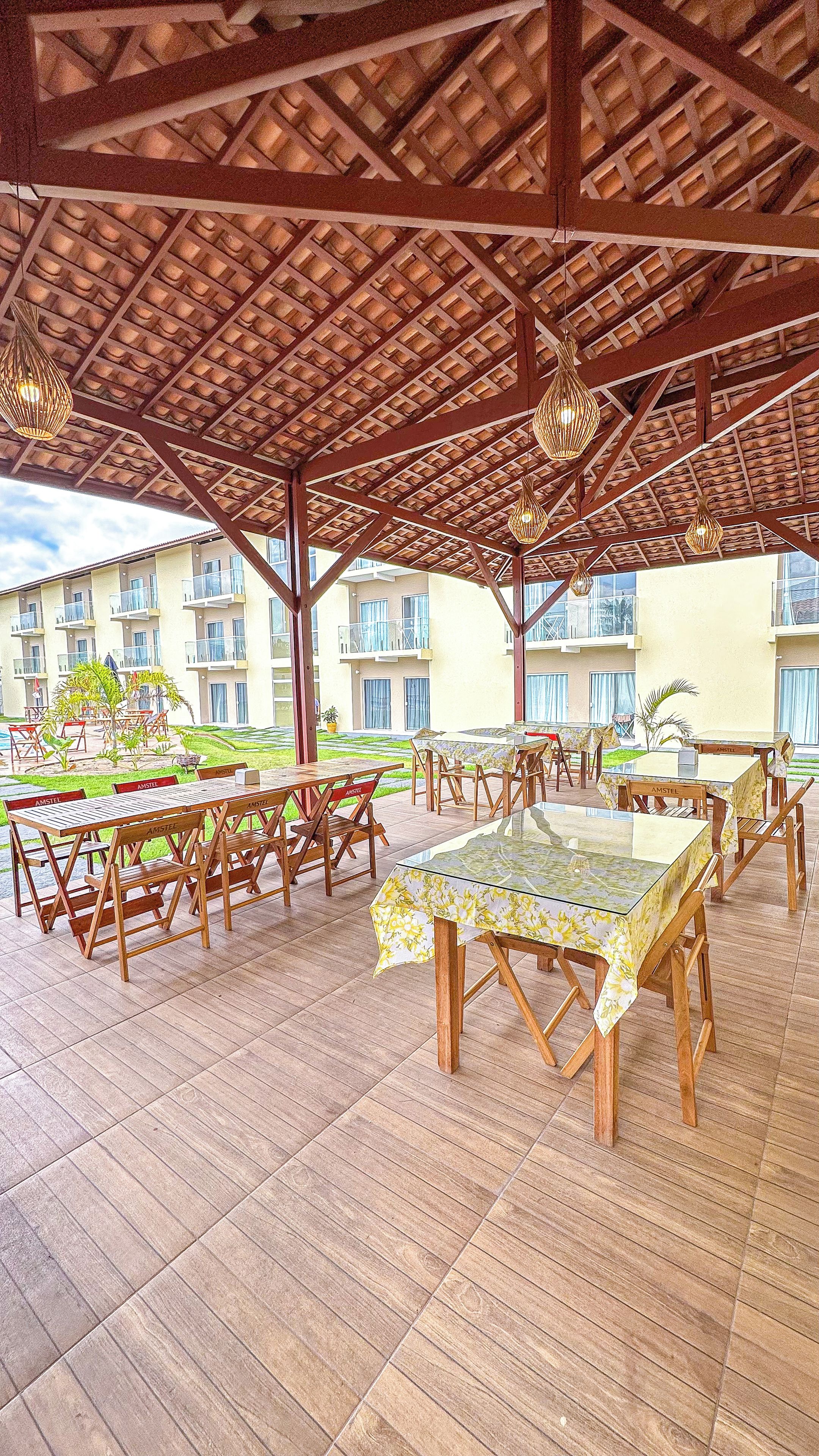 An outdoor patio with wooden tables and chairs, a covered roof structure, and a building with balconies in the background.
