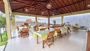 Un área de comedor al aire libre con varias mesas y sillas, piso de baldosas y una estructura de techo de madera.