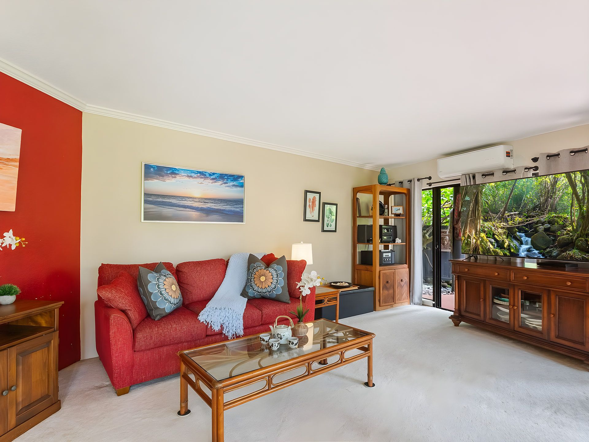 A living room with a red sofa, a glass coffee table, and a view of a natural landscape through a glass door.