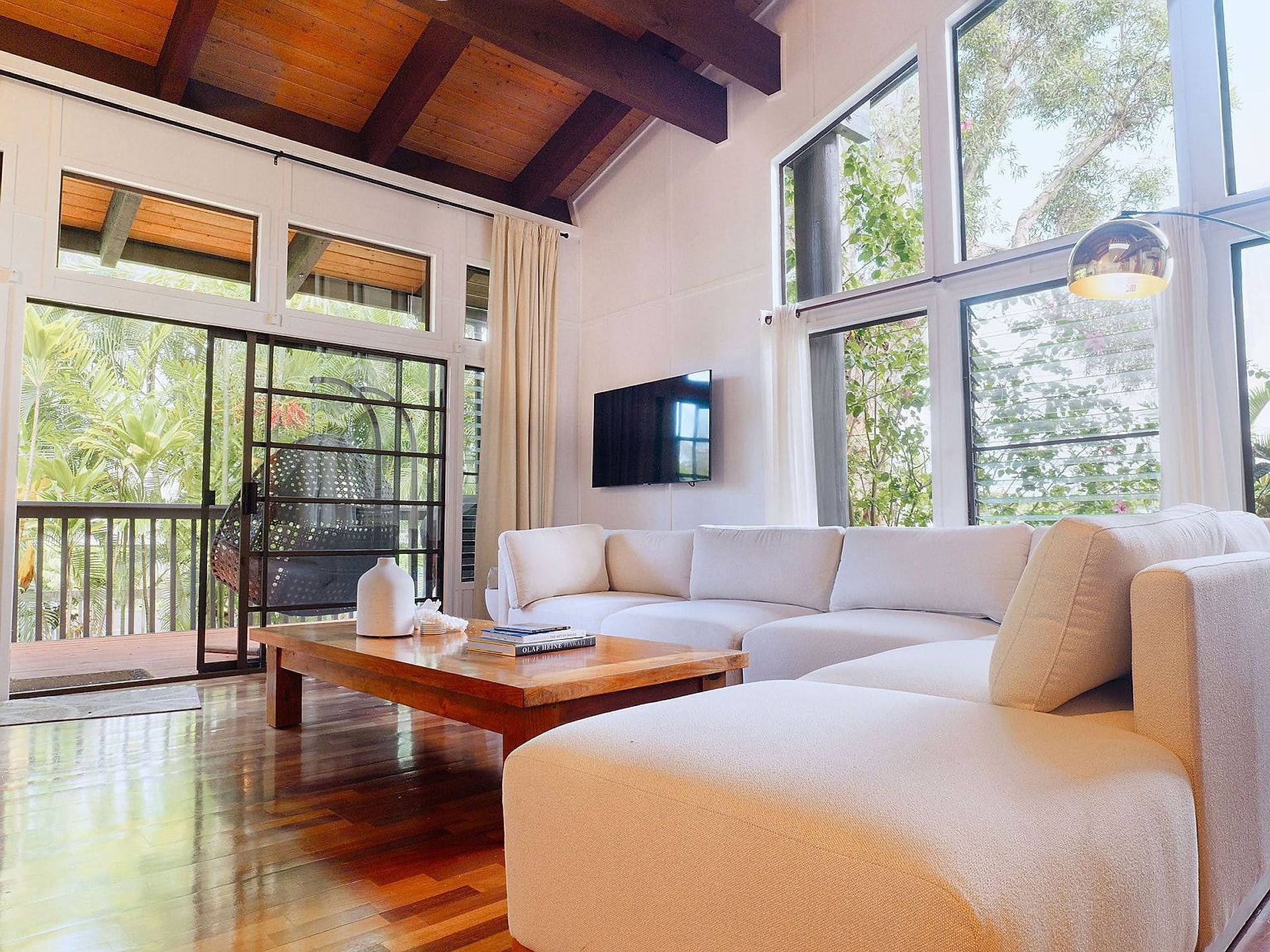A spacious living area with a wooden floor, a white sofa, a coffee table, and a TV mounted on the wall. Large windows provide a view of the outdoors.