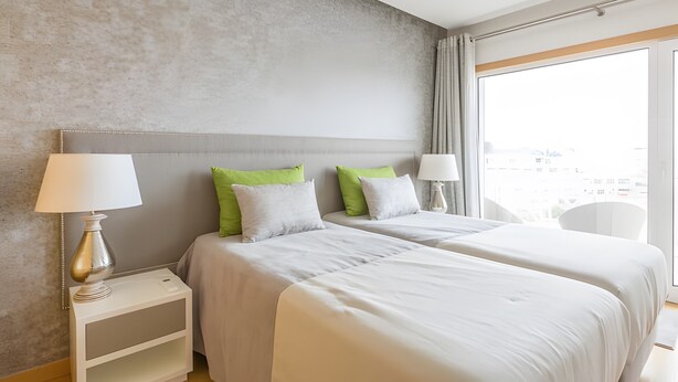 Hotellihuone, jossa on kaksi sänkyä, yöpöytä lampun kanssa, tuoli ja suuri ikkuna, josta on näkymä.