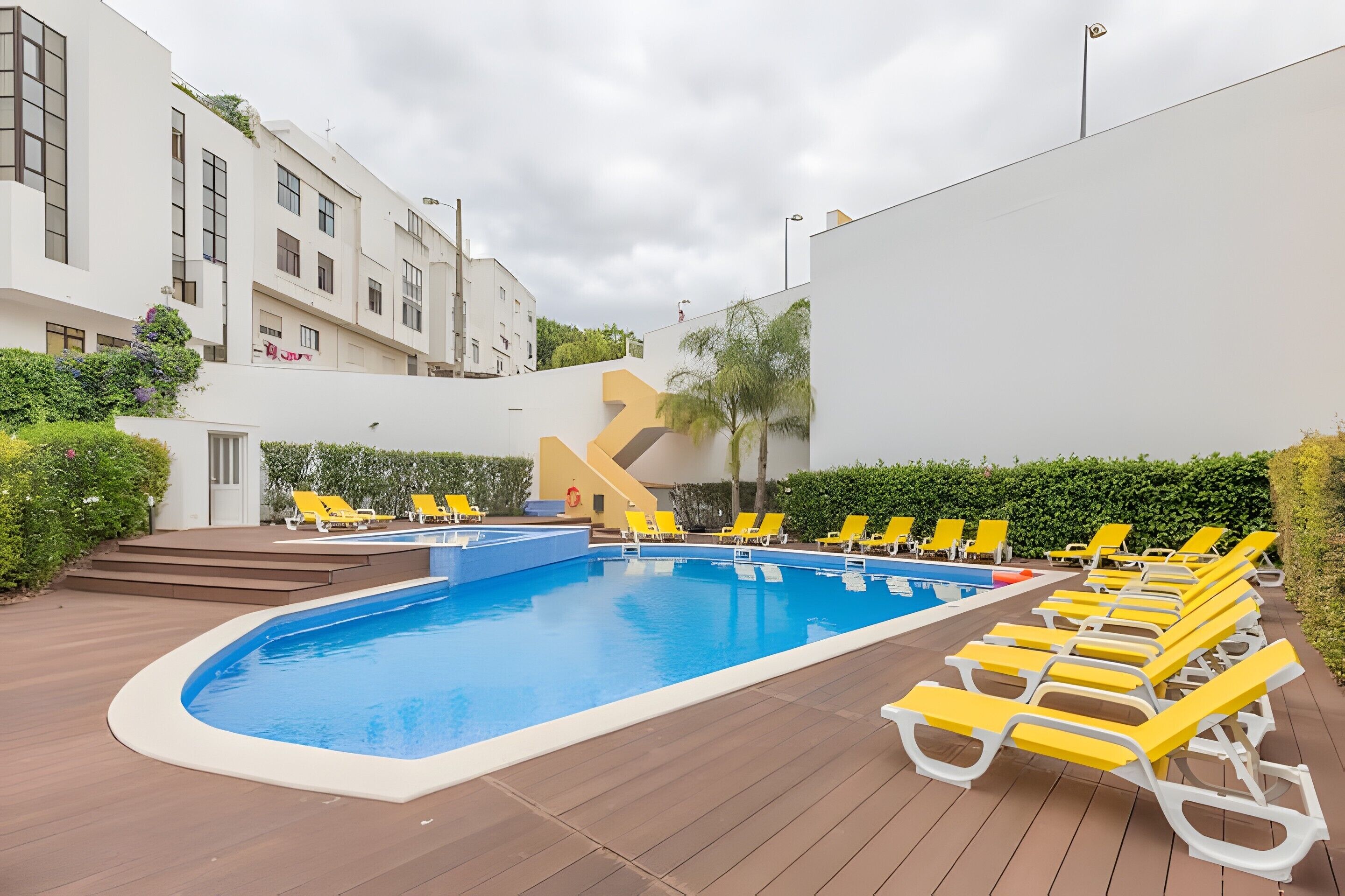 Una piscina con sillas de descanso amarillas, un edificio blanco y un seto.