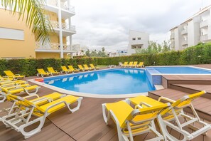 A swimming pool with yellow and white lounge chairs, a wooden deck, and a building with balconies in the background. - 'T1 Garden Hill' Apartment with Shared Pool, Wi-Fi, and Air Conditioning (Albufeira)