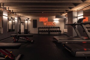 Une salle de sport équipée de tapis de course, d’appareils de musculation et d’un panneau lumineux au néon sur lequel on peut lire : « IRON WORKS ».