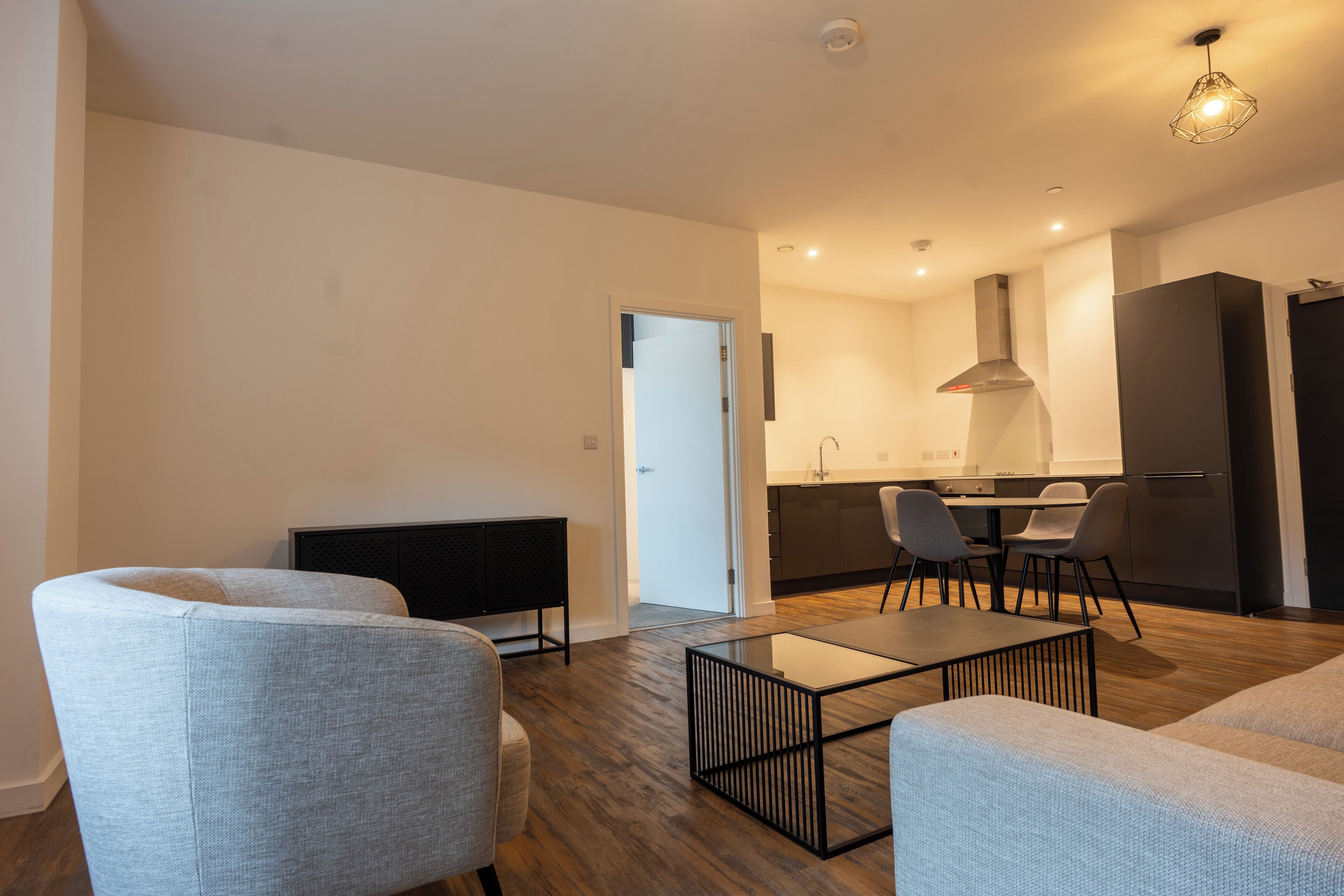 A modern living room with a glass coffee table, two grey armchairs, a dark sofa, and a kitchen area with a dining table and chairs.