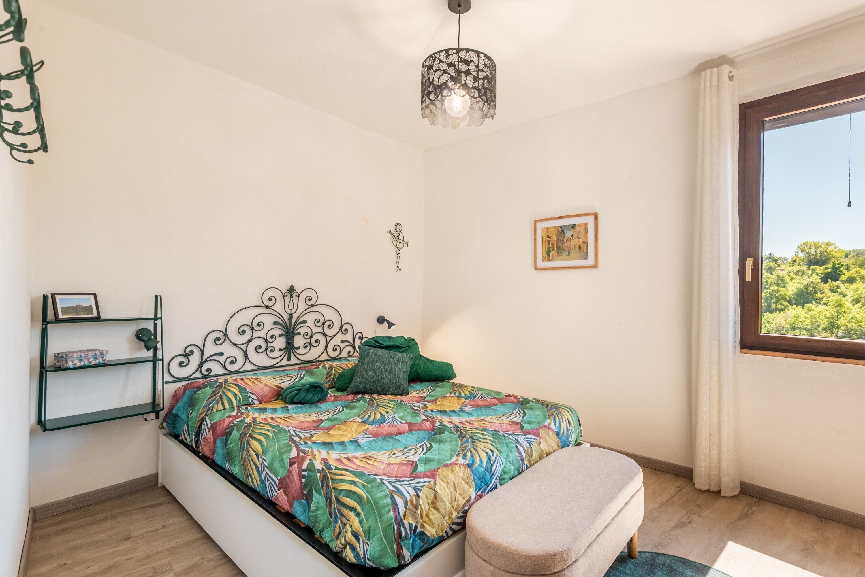 A bedroom with a bed, a bedside table, a hanging light fixture, a window with a view, and a wall-mounted shelf.