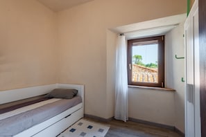 Ein kleines Zimmer mit einem Einzelbett, einem Fenster, einer Tür und einem Lichtschalter an der Wand.