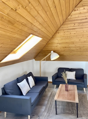Une piÚce de style loft avec un plafond en pente, deux canapés sombres, une table basse en bois et une lucarne.