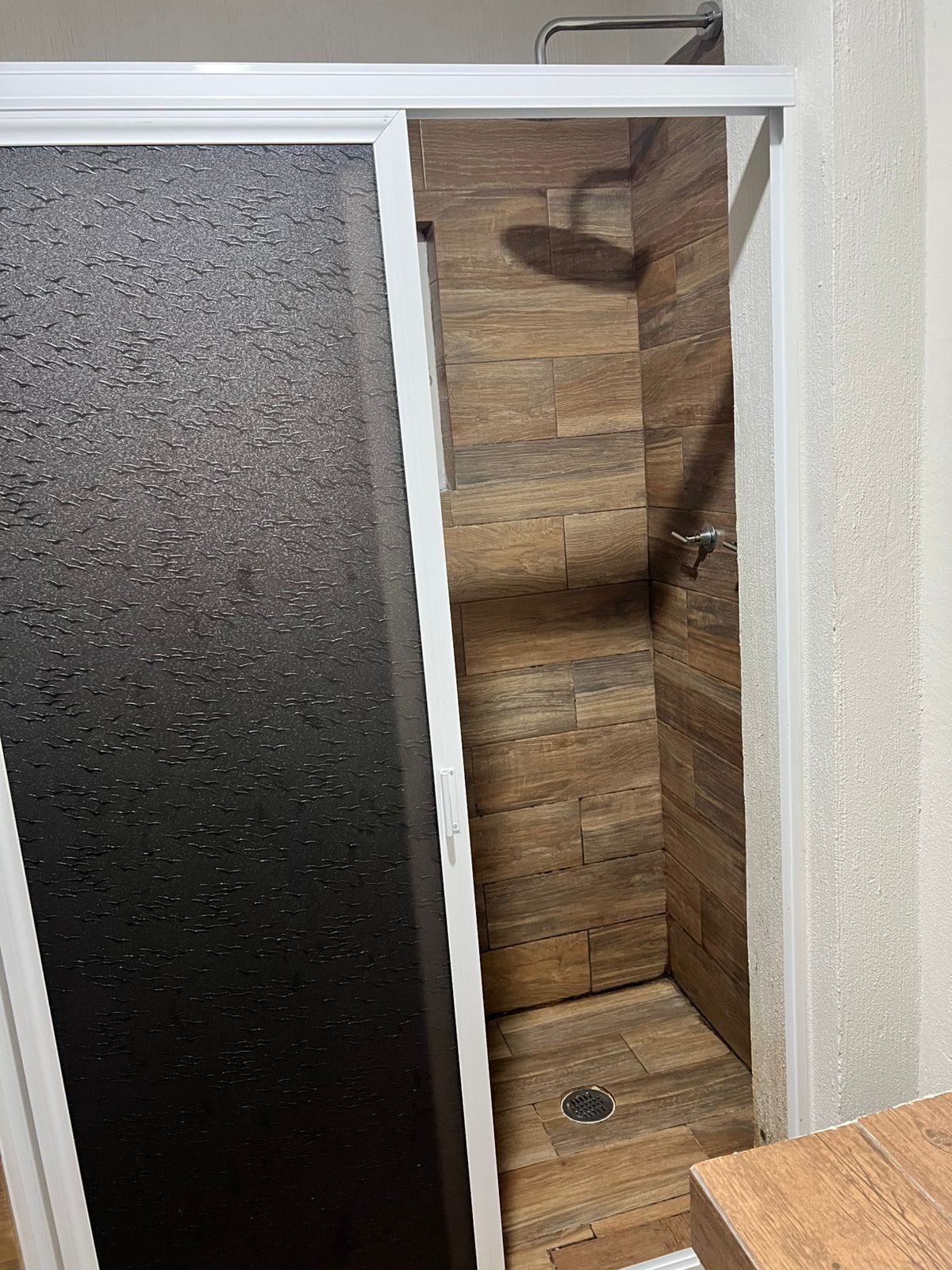 Baño con zona de ducha, paredes de madera y una puerta negra con textura.