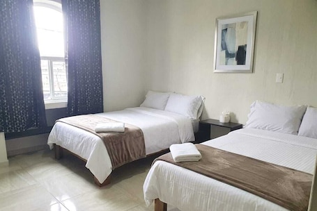 A hotel room with two beds, a window with curtains, a bedside table with a lamp, and a painting on the wall.. Mi Casa Morelia