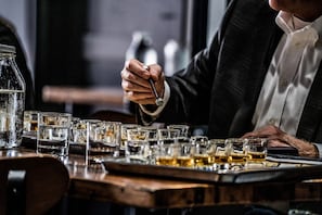 En person hælder drik i et glas i en bar, med flere glas og en vandflaske.
