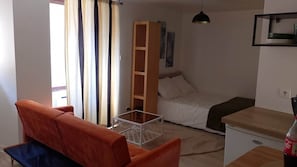 A compact room with a sofa, a bed, a small table, a shelf, and a window with curtains. - Au Pied du Château, the Charm of Heritage (Vizille)