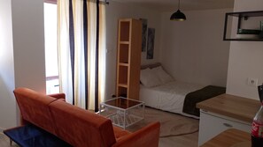 Quarto compacto com cama, uma pequena escrivaninha, sofá e janela com cortinas.