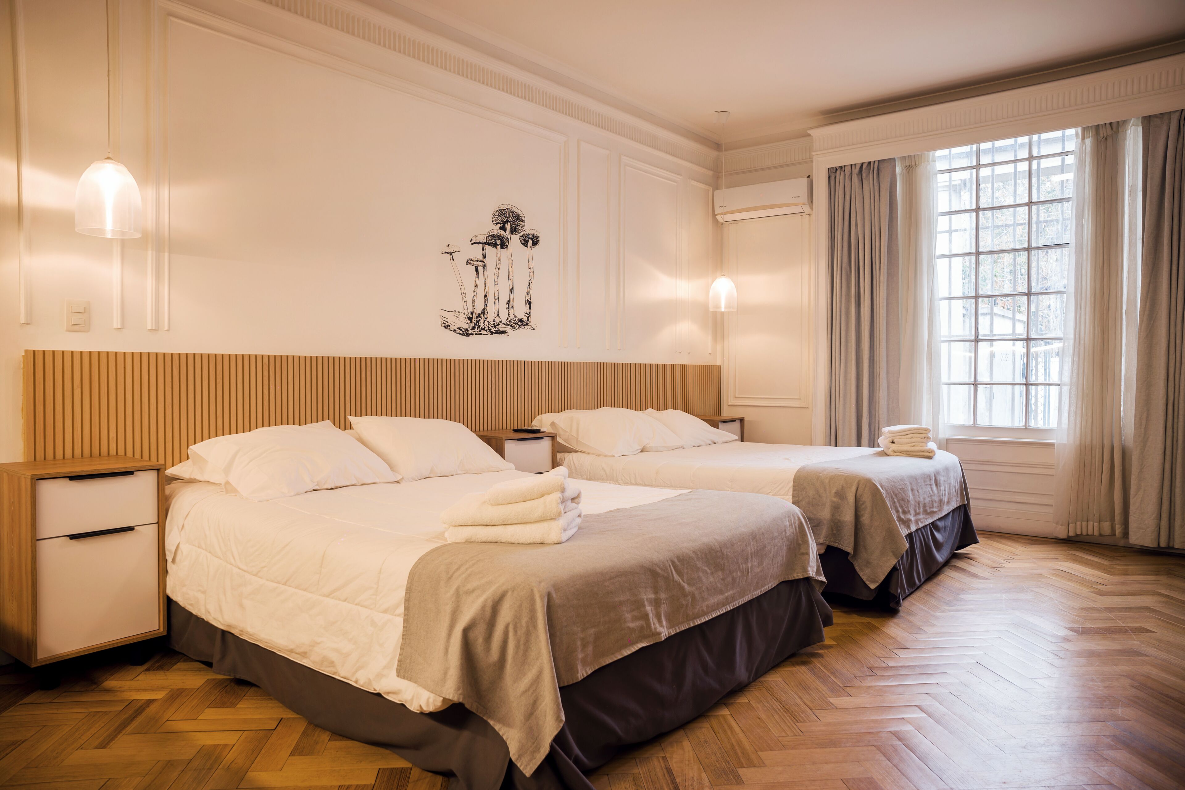 A hotel room with two beds, a wooden floor, a wall-mounted artwork, and a window with curtains.