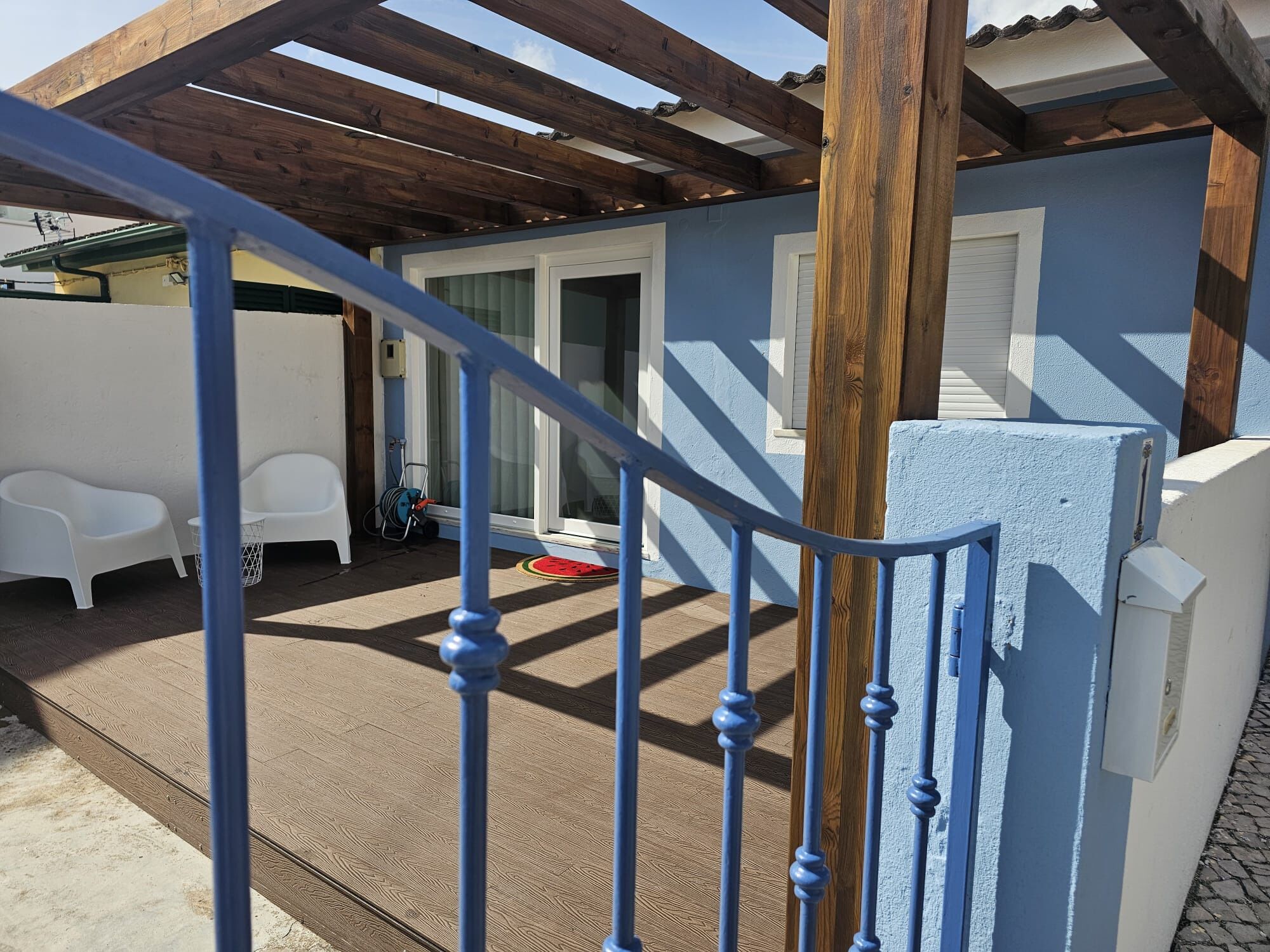 A covered outdoor area with a wooden pergola, a blue railing, and a white door leading to a room.