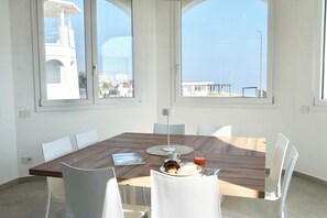 A modern dining room with a large table, white chairs, and a view of a building and sky through large windows.