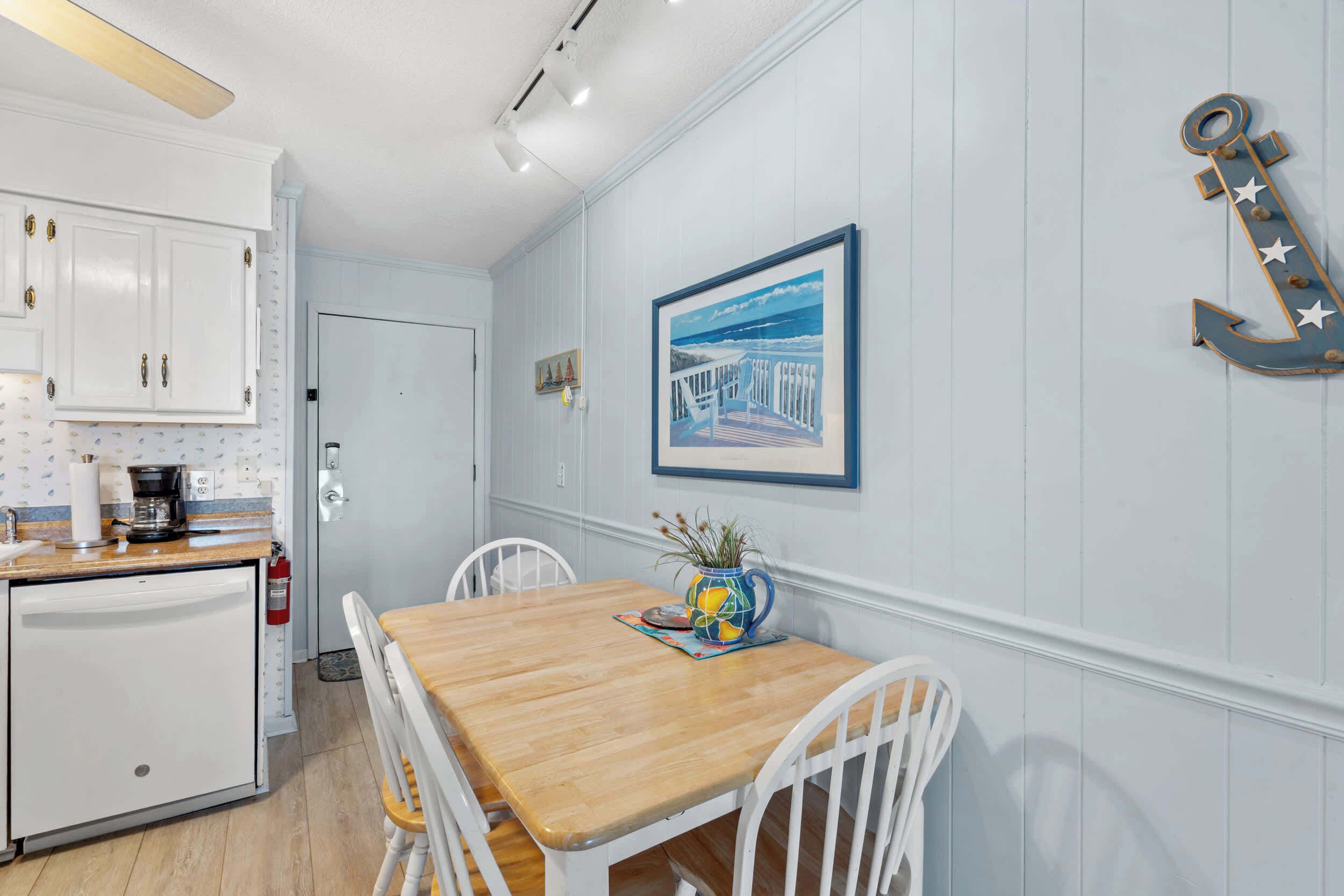 A kitchen with white cabinets, a wooden table, and chairs. There is a framed picture on the wall and a decorative anchor.