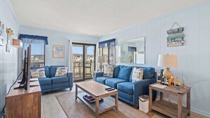 Un salon avec deux canapés bleus, une table basse en bois, une table d’appoint avec une lampe et un panneau décoratif fixé au mur.