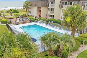 Ein Swimmingpool, umgeben von Liegestühlen und Palmen, mit Blick auf den Ozean und ein Gebäude mit Balkonen.