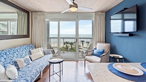 Ein Zimmer mit Deckenventilator, einem an der Wand montierten Fernseher, einer Couch, einem Sessel, einem Tisch und einem Balkon mit Meerblick.