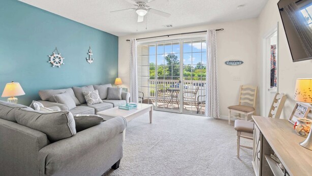 A living room with a teal wall, a ceiling fan, a sofa, a coffee table, and a view through a glass door. - "Seas the Day" at barefoot resort in Ironwood (North Myrtle Beach)