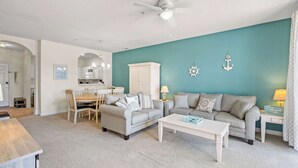 A living room with two sofas, a coffee table, a dining area with a table and chairs, and a kitchen in the background. - "Seas the Day" at barefoot resort in Ironwood (North Myrtle Beach)
