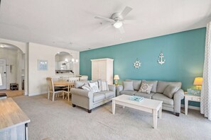 A living room with two sofas, a coffee table, a dining area with a table and chairs, and a kitchen in the background. - "Seas the Day" at barefoot resort in Ironwood (North Myrtle Beach)