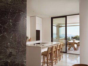 Eine moderne Küche mit Marmor-Spritzschutz, einer zentralen Kochinsel inklusive Barhocker und Blick ins Freie durch eine Schiebetür.