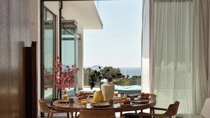 Un comedor con una mesa dispuesta para el desayuno, un jarrón con flores y vistas al mar a través de grandes ventanales.