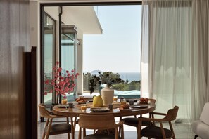 Ein Essbereich mit einem für das Frühstück gedeckten Tisch, einer Blumenvase und Meerblick durch große Fenster.