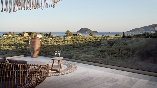 Habitación en un rascacielos con un ventanal grande que da a un paisaje costero, una silla de mimbre, una mesa redonda de madera con dos vasos y un jarrón grande.