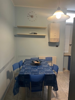 Ein Essbereich mit einem Tisch für vier Personen, blauen Stühlen, einer Wanduhr und einer Deckenleuchte.