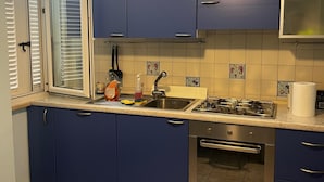 Keittiössä on siniset kaapit, kaasuliesi, pesuallas ja ikkuna, jossa on sälekaihtimet.