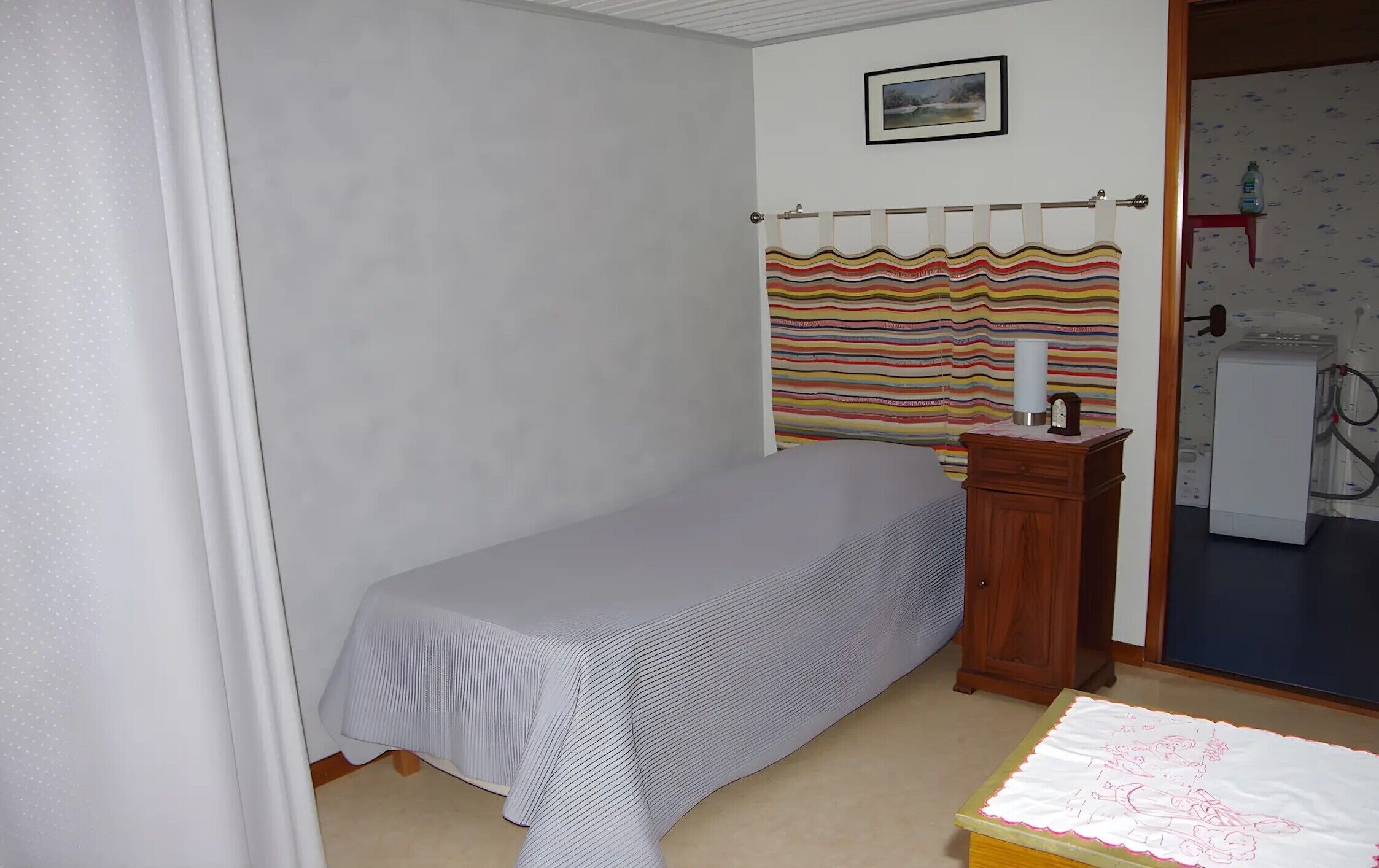 Ein kleines Zimmer mit einem Einzelbett, einem Holznachttisch, einer Nachttischlampe, einem Streifenmuster-Vorhang und einem an der Wand hängenden Bild.