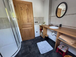 Ein Badezimmer mit Holztür, Waschbecken, Spiegel, Dusche und einem kleinen Hocker.