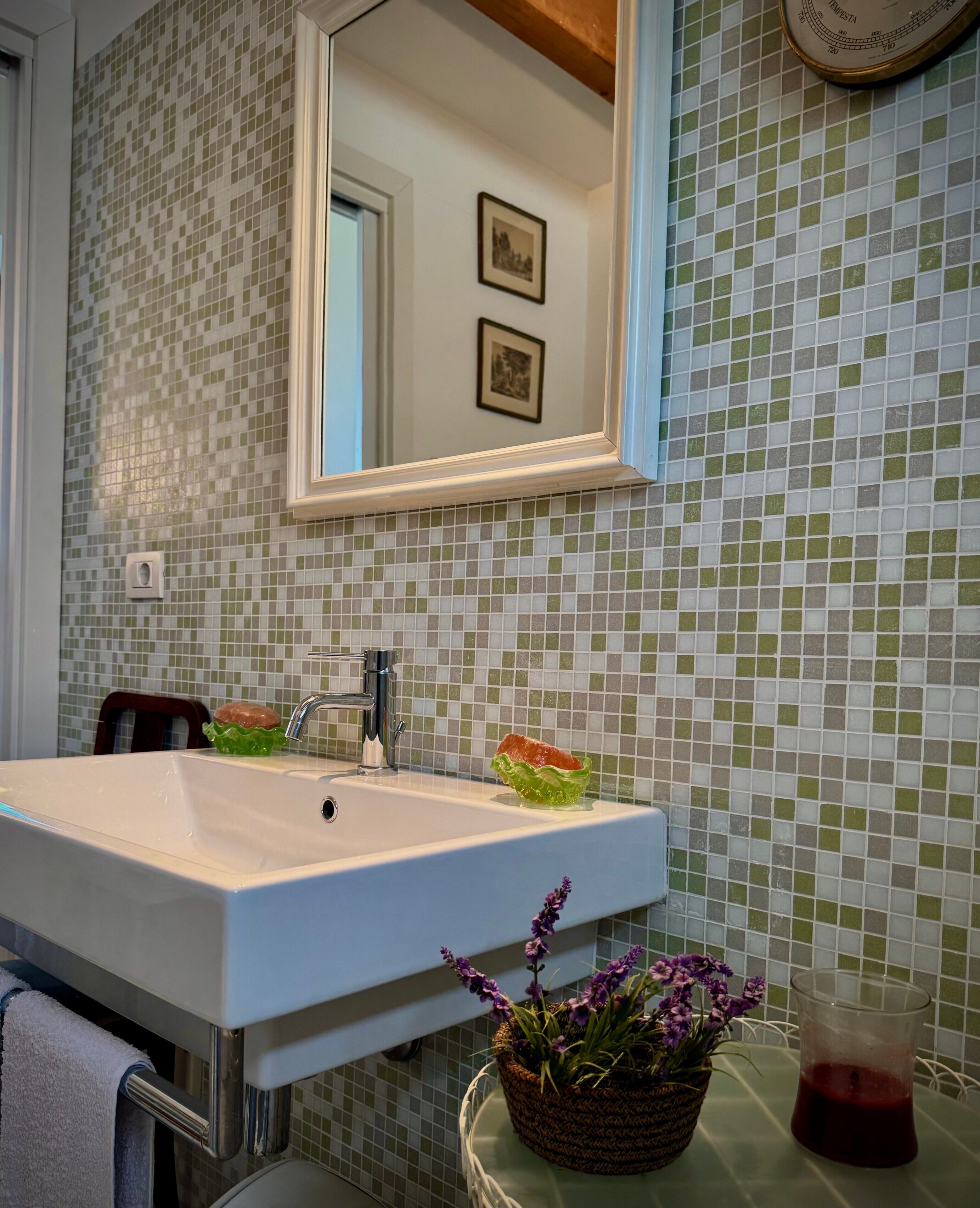 Une salle de bain avec un lavabo blanc, un miroir, un mur recouvert de carreaux verts et blancs, une horloge et une plante en pot.