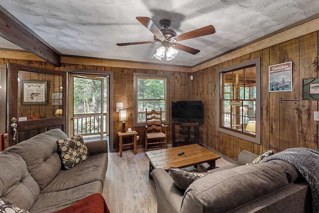 A cabin-style room with a wooden interior, a ceiling fan, a television, and a large window.