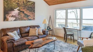 A living room with a brown leather sofa, a coffee table, a painting of a river, a tripod lamp, and a view through large windows.