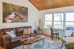 A living room with a brown leather sofa, a coffee table, a painting of a river, a tripod lamp, and a view through large windows. - Elevated mountain-view cabin with private hot tub, 2 decks, firepit, & grill (Cullowhee)