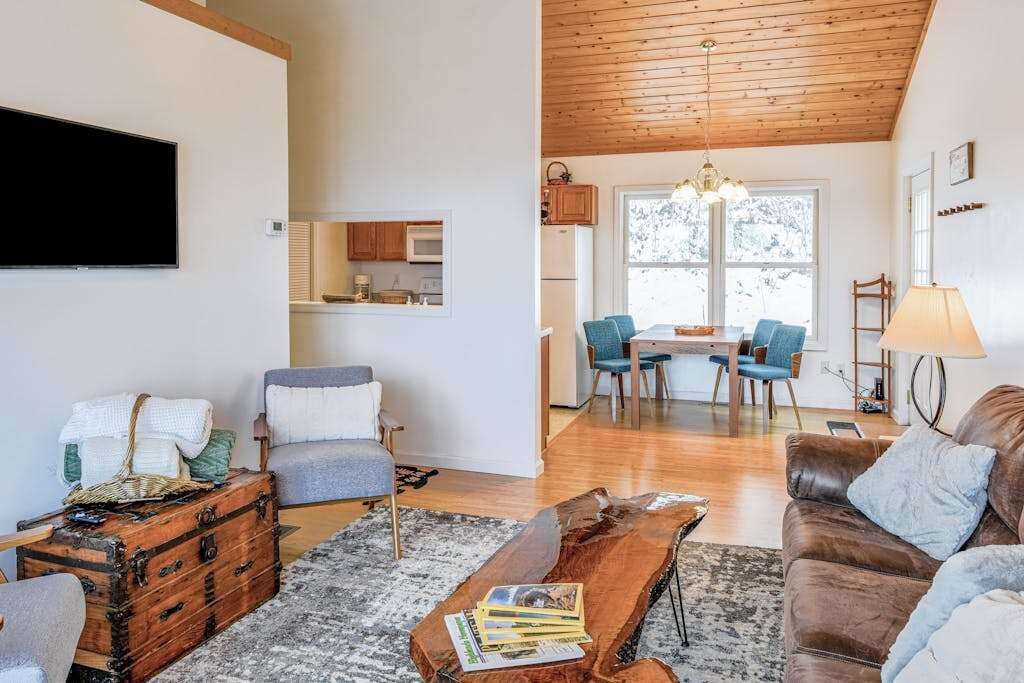 A living room with a wooden ceiling, a TV mounted on the wall, a dining area with a table and chairs, and a kitchen in the background.