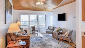 A modern living room with a wooden ceiling, a large window, a flat-screen TV, a sofa, a coffee table, and a fireplace.