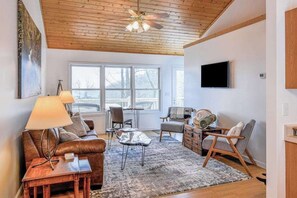A modern living room with a wooden ceiling, a large window, a flat-screen TV, a sofa, a coffee table, and a fireplace. - Elevated mountain-view cabin with private hot tub, 2 decks, firepit, & grill (Cullowhee)
