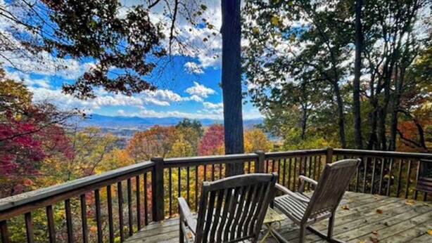 Une terrasse en bois avec deux chaises en bois, donnant sur une forêt aux couleurs automnales et sur une chaîne de montagnes lointaine.