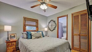A bedroom with a bed, a ceiling fan, a window with blinds, a wall clock, and a wooden door.