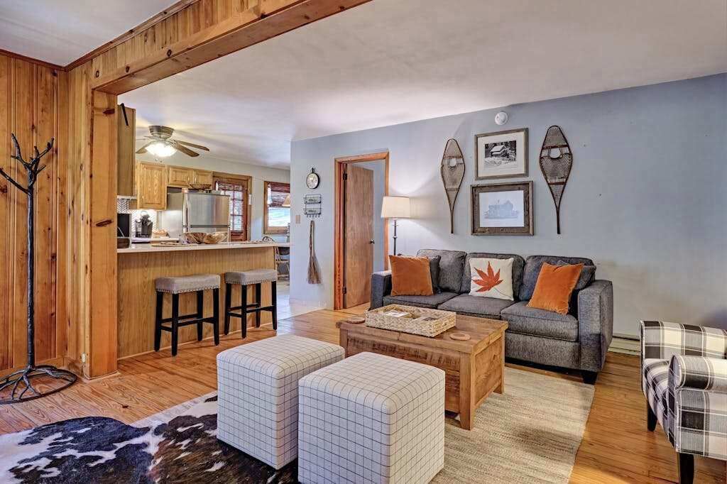 A cozy living room with a sofa, ottomans, and a coffee table. A kitchen is visible in the background with wooden cabinets and a bar stool.